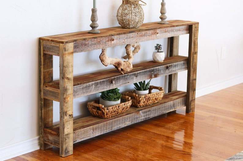 Puede incluir: Mesa consola de madera r&uacute;stica con tres estantes. La mesa est&aacute; hecha de madera recuperada y tiene un acabado natural. Cuenta con una pieza decorativa de madera a la deriva en el estante superior y dos plantas en maceta en el estante inferior.