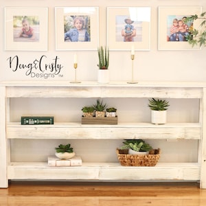 May include: Whitewashed wooden console table with three shelves. The table has a distressed white finish and is decorated with plants and framed photos. The text "Doug & Christy Designs™" is visible on the wall above the table.