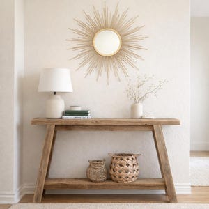 May include: A rustic wooden console table with a lower shelf, holding two woven baskets. A white ceramic lamp and books sit on the table. Above, a gold sunburst mirror hangs on the textured wall. A vase with greenery is also on the table.