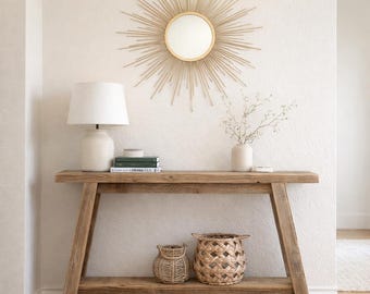 May include: A rustic wooden console table with a lower shelf, holding two woven baskets. A white ceramic lamp and books sit on the table. Above, a gold sunburst mirror hangs on the textured wall. A vase with greenery is also on the table.