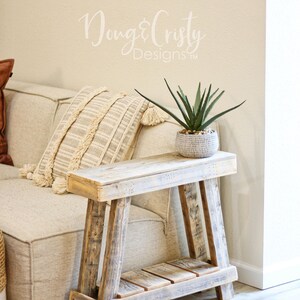 May include: A rustic wooden side table with a whitewashed finish and a lower shelf. The table is next to a white couch with a brown throw pillow and a white pillow with fringe. A potted green plant sits on the table.