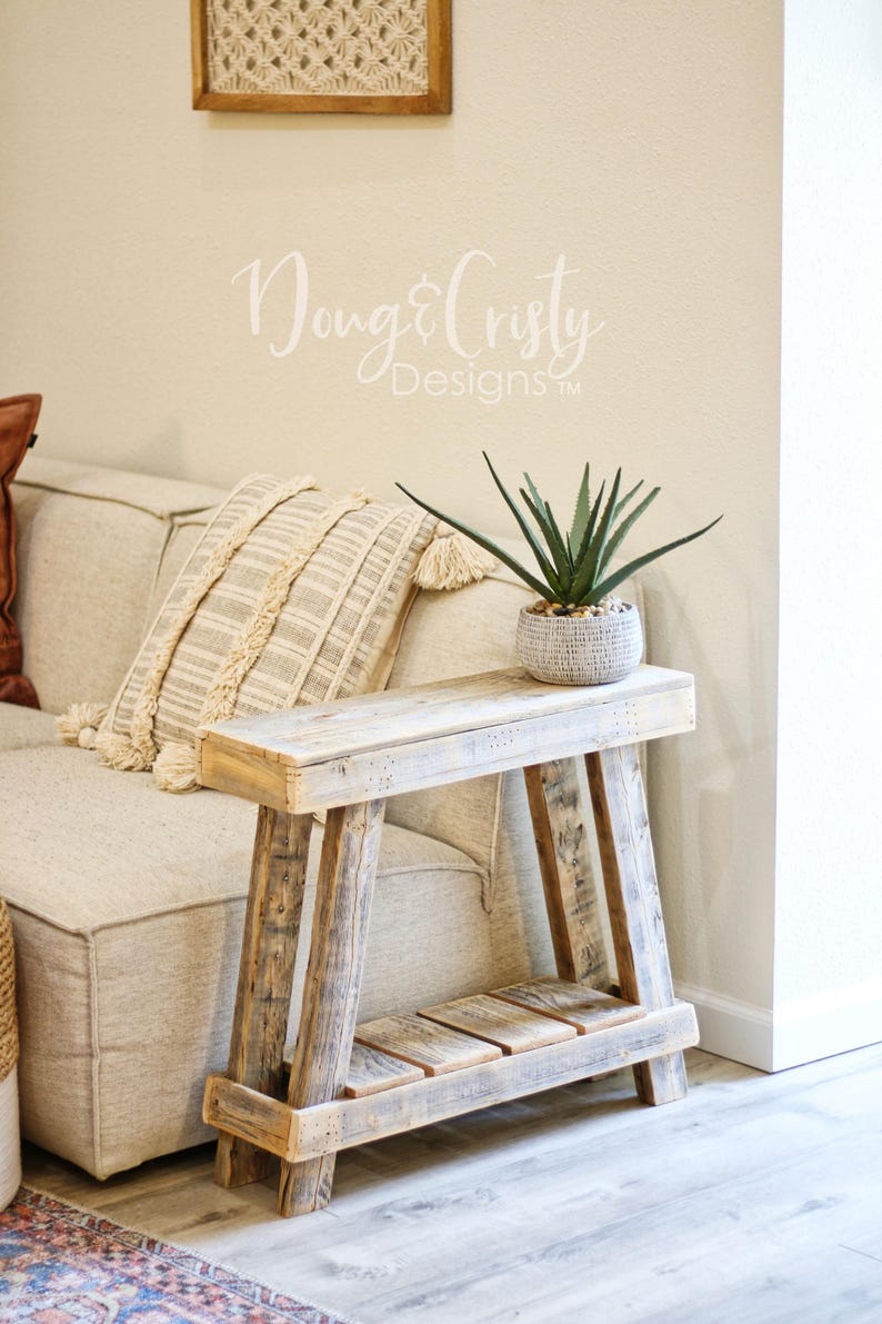 May include: A rustic wooden side table with a whitewashed finish and a lower shelf. The table is next to a white couch with a brown throw pillow and a white pillow with fringe. A potted green plant sits on the table.