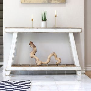 May include: A white wooden console table with a distressed finish. The table has a dark wood top and a lower shelf. A decorative piece of driftwood sits on the shelf. Two gold candle holders and a potted plant are on the table.