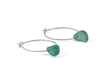 May include: A pair of silver hoop earrings with a teal blue sea glass charm on each hoop.