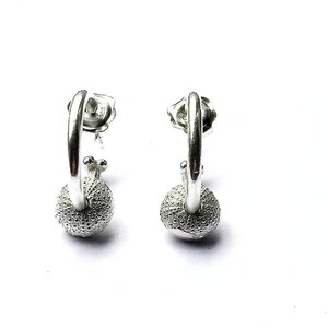 Pode incluir: Um par de brincos de argola de prata com um design de bola texturizada. Os brincos são feitos de metal e têm um design simples e elegante.