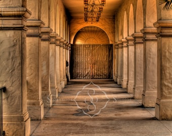 May include: A long, arched hallway with white columns and a wrought iron chandelier hanging from the ceiling. The hallway is lit by sunlight streaming in from the windows at the end of the hallway.