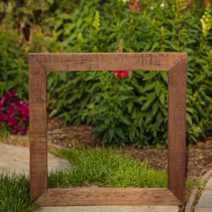 May include: A rustic wooden frame with a natural wood finish. The frame is standing upright in a grassy area with a blurred background of green foliage and red flowers.