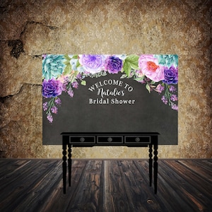 May include: A black chalkboard with purple and green floral decorations and the text "Welcome to Natalie's Bridal Shower". A black table with three drawers is in front of the chalkboard.