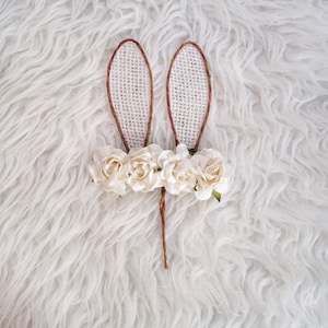 May include: A pair of brown bunny ears made of burlap with white fabric flowers attached to the base.