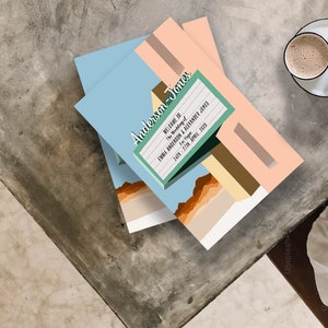 May include: A stack of light blue, peach, and brown invitation cards with a retro-style sign that reads "Welcome to the Wedding of Emma Anderson & Alexander Jones. Save the Date. Saturday, 27th April 2023."