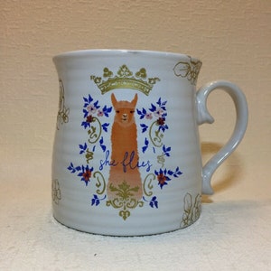 May include: White ceramic mug with a gold crown, floral design, and a llama with the text "she flies" in blue.