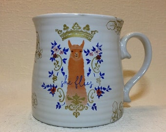 May include: White ceramic mug with a gold crown, floral design, and a llama with the text "she flies" in blue.