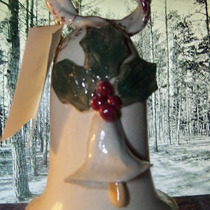 May include: A ceramic Christmas bell with a twisted handle, decorated with holly leaves and red berries. The bell has a smaller bell-shaped clapper and a cream-colored finish with red accents. A blank tag hangs from the handle.