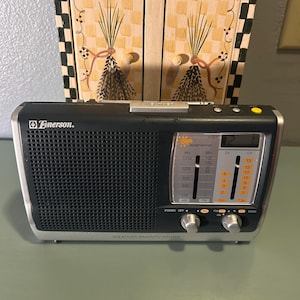 May include: Black Emerson weather band radio with silver trim. Features a speaker grill, tuning dials, and a digital display. The radio has the text "WEATHER BAND/TV SOUND" and "Emerson."
