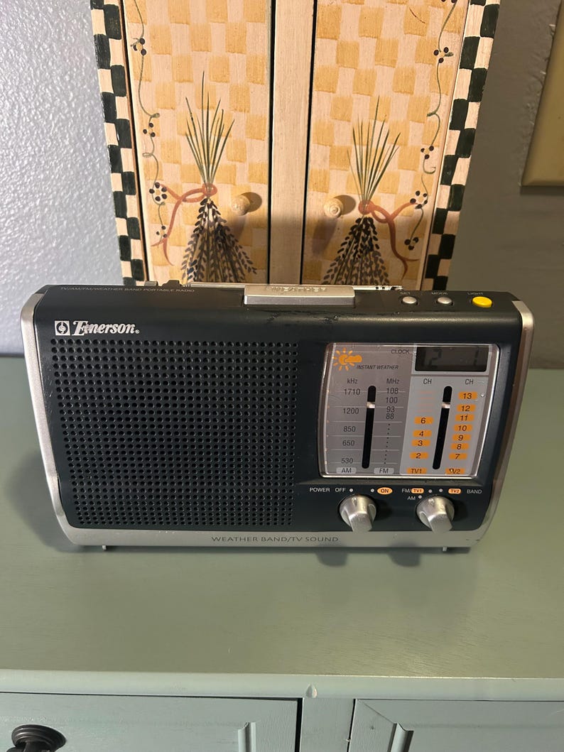 May include: Black Emerson weather band radio with silver trim. Features a speaker grill, tuning dials, and a digital display. The radio has the text "WEATHER BAND/TV SOUND" and "Emerson."