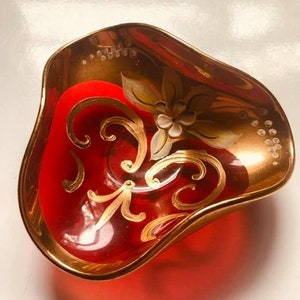 Puede incluir: Un cuenco de vidrio rojo con diseños florales pintados en oro y blanco. El cuenco tiene una forma ondulada y orgánica.