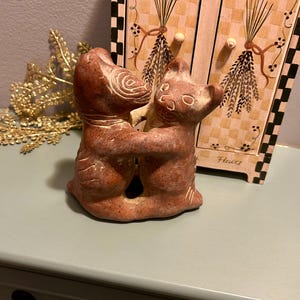 May include: A terracotta-colored sculpture of two embracing cat figures. The cats are stylized with etched details and a rough texture. The sculpture is on a light green surface, with a decorative cabinet and gold-colored foliage in the background.