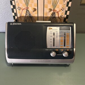 May include: A vintage Emerson radio with a black speaker grill and silver trim. The radio has a tuning dial, power and band selection knobs, and the text "WEATHER BAND/TV SOUND". The radio is mounted on a green surface.