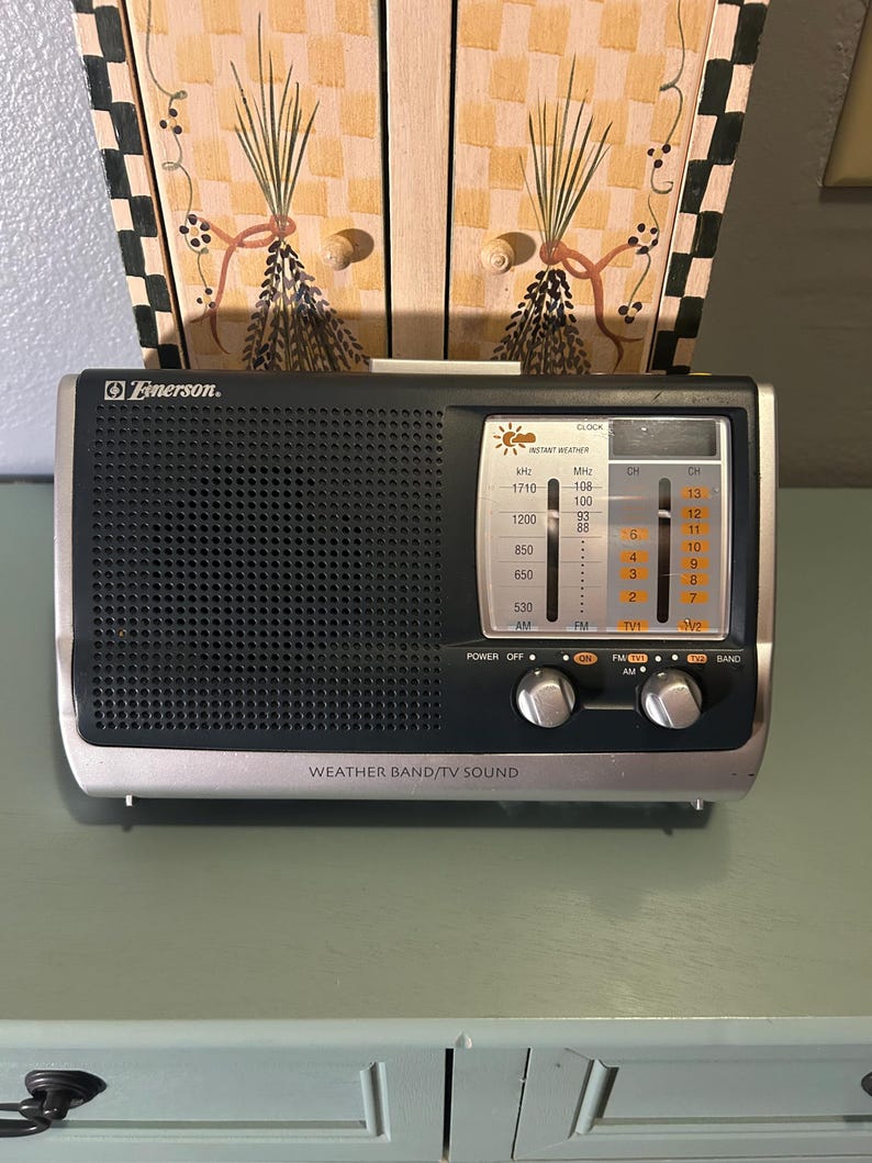 May include: A vintage Emerson radio with a black speaker grill and silver trim. The radio has a tuning dial, power and band selection knobs, and the text "WEATHER BAND/TV SOUND". The radio is mounted on a green surface.