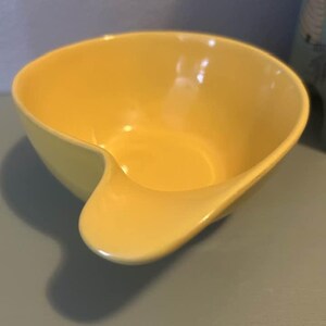 May include: A bright yellow ceramic bowl with a unique, curved handle. The bowl has a smooth, glossy finish and a rounded shape. The design is modern and minimalist, suitable for serving food or as a decorative piece.