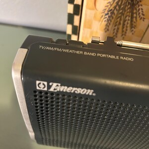 May include: A close-up of a black Emerson portable radio. The radio has the text "TV/AM/FM/WEATHER BAND PORTABLE RADIO" printed on the top. The Emerson logo is visible on the front of the radio.