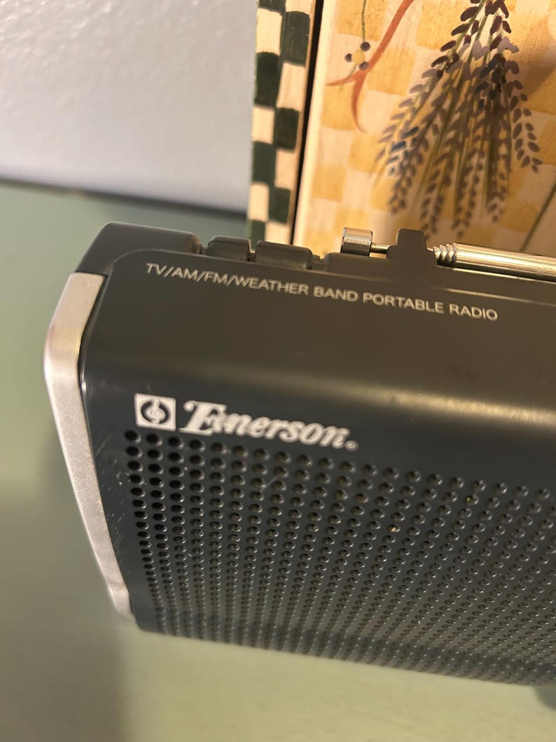 May include: A close-up of a black Emerson portable radio. The radio has the text "TV/AM/FM/WEATHER BAND PORTABLE RADIO" printed on the top. The Emerson logo is visible on the front of the radio.