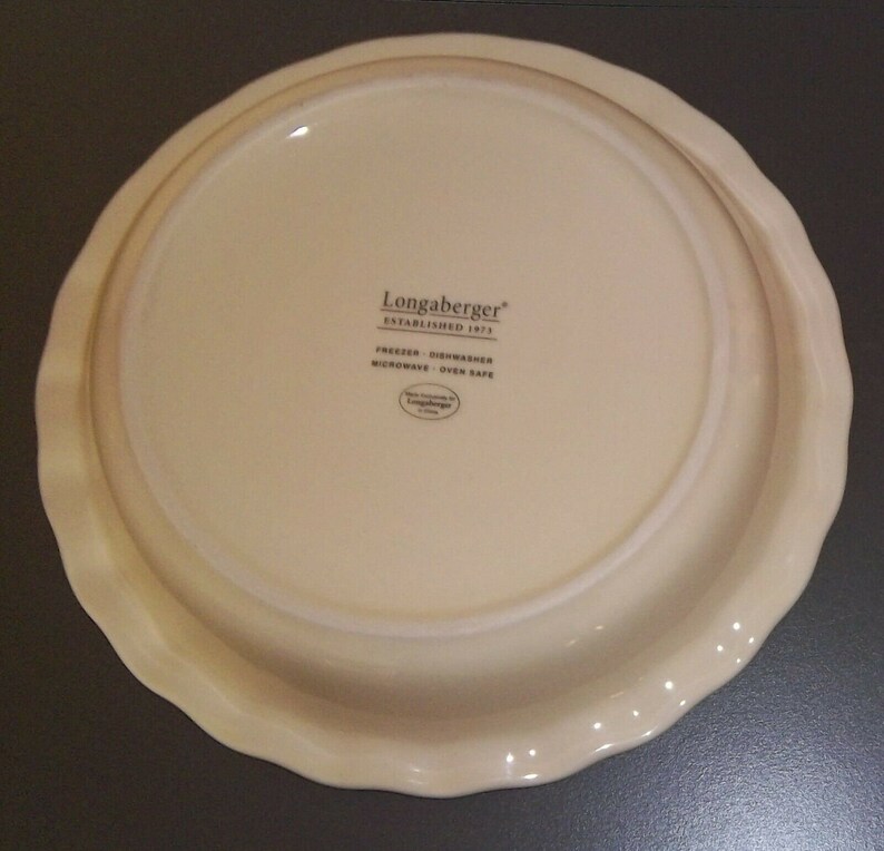 May include: Off-white pie plate with a scalloped edge. The bottom of the plate has the text "Longaberger ESTABLISHED 1973" and "FREEZER - DISHWASHER - MICROWAVE - OVEN SAFE".
