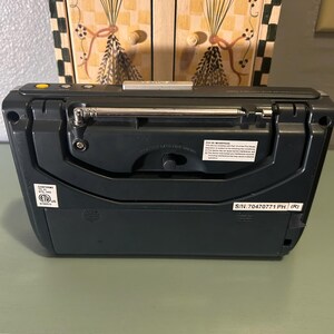 May include: A dark gray portable radio with a retractable antenna. The radio has a small dial and several buttons. The back of the radio has a label with text and a serial number. The radio is sitting on a light green surface.