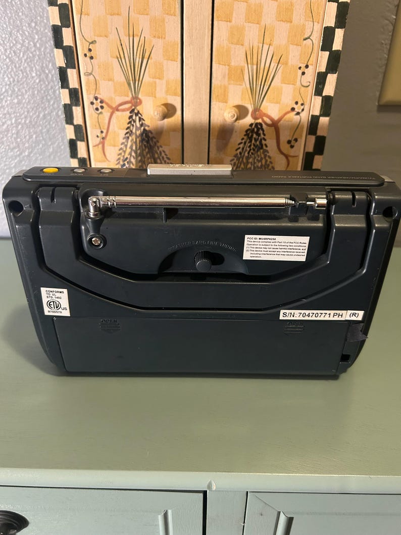 May include: A dark gray portable radio with a retractable antenna. The radio has a small dial and several buttons. The back of the radio has a label with text and a serial number. The radio is sitting on a light green surface.