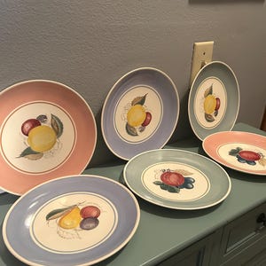 May include: A collection of vintage ceramic plates with fruit designs. The plates feature a variety of colors, including pink, blue, and green, with hand-painted illustrations of lemons, plums, and other fruits. The plates are arranged on a blue cabinet.
