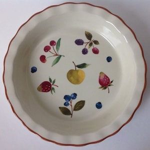 May include: Off-white ceramic pie dish with a scalloped edge and a brown rim. The interior features hand-painted illustrations of various fruits, including cherries, blackberries, an apple, strawberries, and blueberries. A decorative kitchenware item.