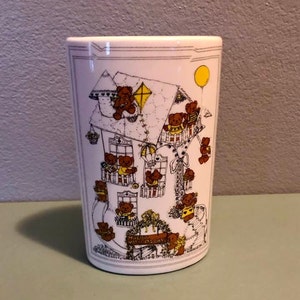 May include: White ceramic mug with a whimsical illustration of a house with teddy bears on the balconies and the words "Merry Mother's Day" on a sign. The house is surrounded by teddy bears and a yellow balloon.
