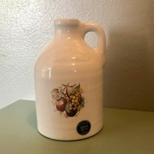 May include: White ceramic pitcher with a handle, featuring a floral design with fruit and a green leaf. The pitcher has a sticker on the bottom that says "Made in the USA Bakers Ridge Chicago".