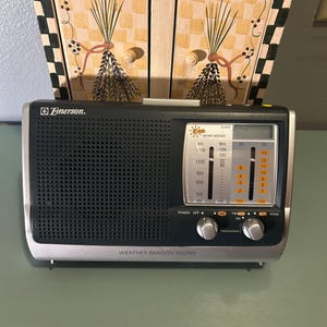 May include: A silver and black Emerson radio with a digital display and tuning knobs. The radio has the words "WEATHER BAND/TV SOUND" printed on the front. The radio is sitting on a green surface.