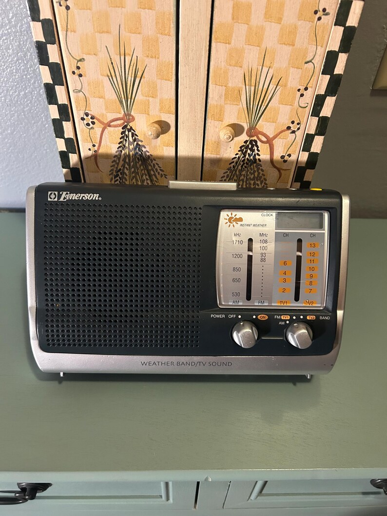 May include: A silver and black Emerson radio with a digital display and tuning knobs. The radio has the words "WEATHER BAND/TV SOUND" printed on the front. The radio is sitting on a green surface.