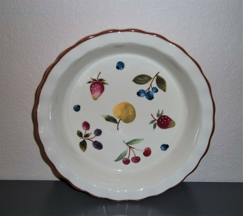 May include: White ceramic pie plate with a scalloped edge and a brown rim. The plate is decorated with colorful illustrations of various fruits, including strawberries, blueberries, cherries, and a lemon.