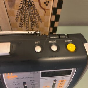May include: A black and silver portable radio with a retractable antenna. The radio has buttons labeled "SET", "MODE", and "LIGHT". The display shows frequency and channel information. The text "INSTANT WEATHER" is visible on the radio.