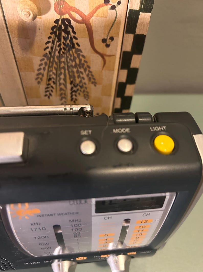 May include: A black and silver portable radio with a retractable antenna. The radio has buttons labeled "SET", "MODE", and "LIGHT". The display shows frequency and channel information. The text "INSTANT WEATHER" is visible on the radio.