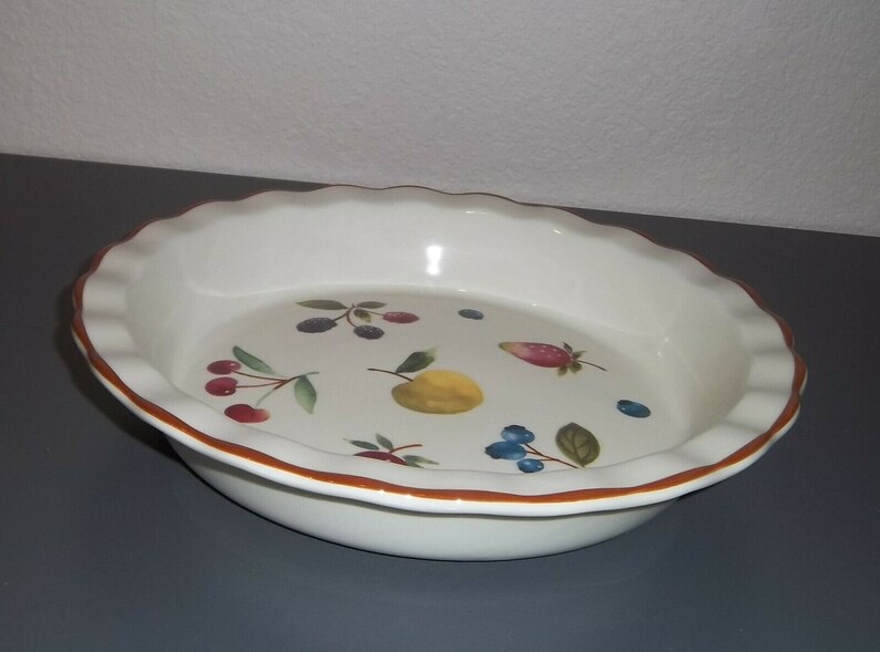 May include: White ceramic pie dish with a scalloped edge and a brown rim. The interior features a colorful fruit design, including cherries, blueberries, a lemon, and a strawberry. A decorative and functional kitchenware item.