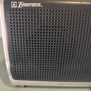 May include: Close-up of a vintage Emerson weather band radio. The radio has a dark gray perforated speaker grill with the Emerson logo in white. The bottom of the radio is silver with the text "WEATHER BAND/TV SOUND".