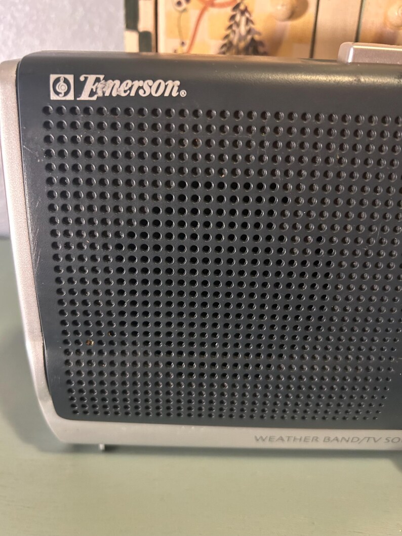 May include: Close-up of a vintage Emerson weather band radio. The radio has a dark gray perforated speaker grill with the Emerson logo in white. The bottom of the radio is silver with the text "WEATHER BAND/TV SOUND".