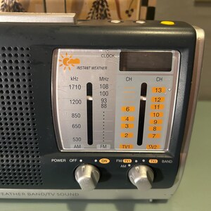 May include: A black and silver portable radio with the words "Instant Weather" and "Weather Band/TV Sound" printed on the front. It has a digital clock display and several dials and buttons for power, tuning, and channel selection.