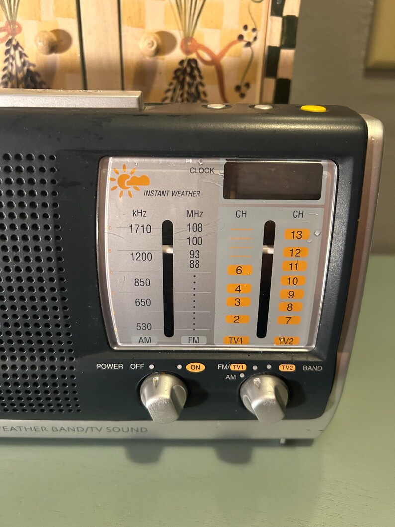 May include: A black and silver portable radio with the words "Instant Weather" and "Weather Band/TV Sound" printed on the front. It has a digital clock display and several dials and buttons for power, tuning, and channel selection.
