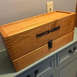 May include: A wooden tool box with the words "PORTER+CABLE" printed in black. The box is made of light-colored wood and has a metal latch. The box is rectangular and sits on a blue cabinet.