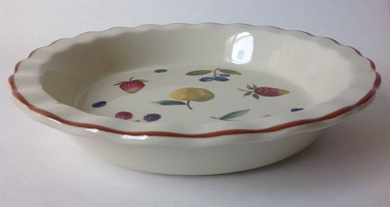 May include: Off-white ceramic pie dish with a scalloped edge and a brown rim. The interior features painted illustrations of strawberries, blueberries, and a yellow fruit. A decorative kitchenware item.