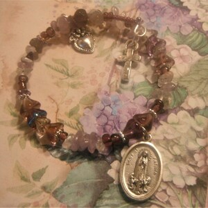 May include: A purple and brown beaded bracelet with a silver charm of the Virgin Mary, a heart, and a cross. The bracelet is on a floral patterned background.
