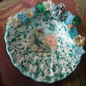 May include: A decorative seashell dish, embellished with glitter, faux flowers, and a mermaid charm. The dish features a central image of a mermaid with an umbrella. The edges are trimmed with blue and green accents, creating a whimsical, ocean-themed aesthetic.