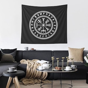 May include: A black and white tapestry with a white compass design surrounded by runes. The tapestry is hanging on a white wall above a gray couch with pillows and a throw blanket.
