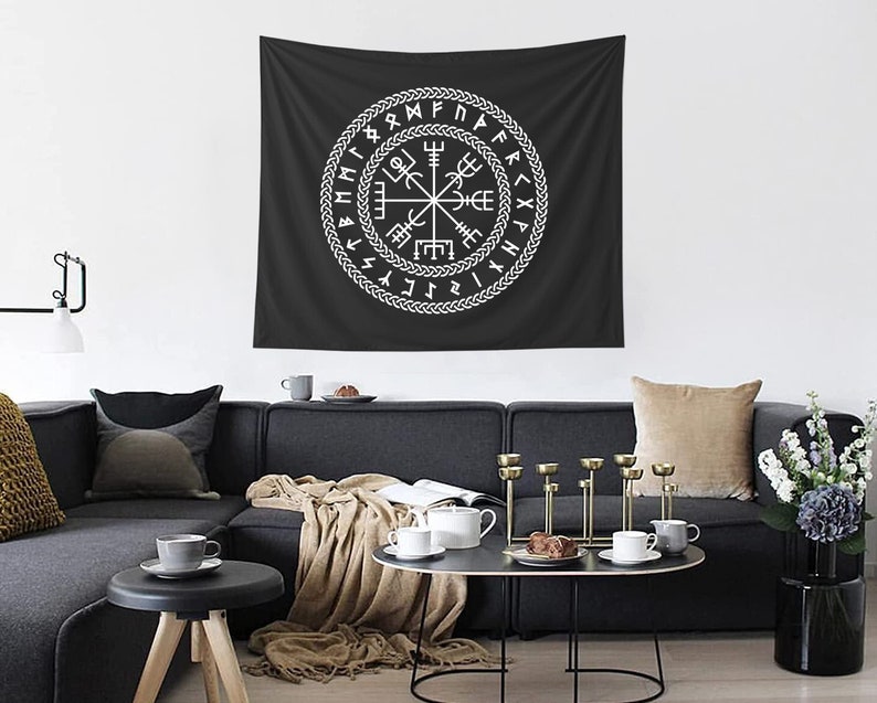 May include: A black and white tapestry with a white compass design surrounded by runes. The tapestry is hanging on a white wall above a gray couch with pillows and a throw blanket.