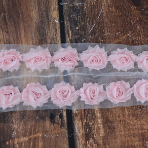 May include: Pink fabric rose trim on a white mesh background. The trim is made of multiple layers of fabric to create a 3D effect.